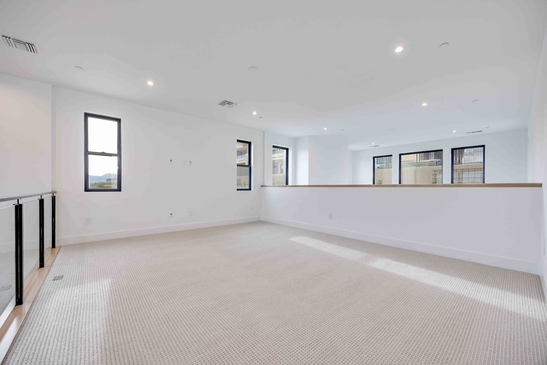 A spacious, well-lit room with a clean, minimalist design featuring white walls, a carpeted floor, and large windows providing natural light.
