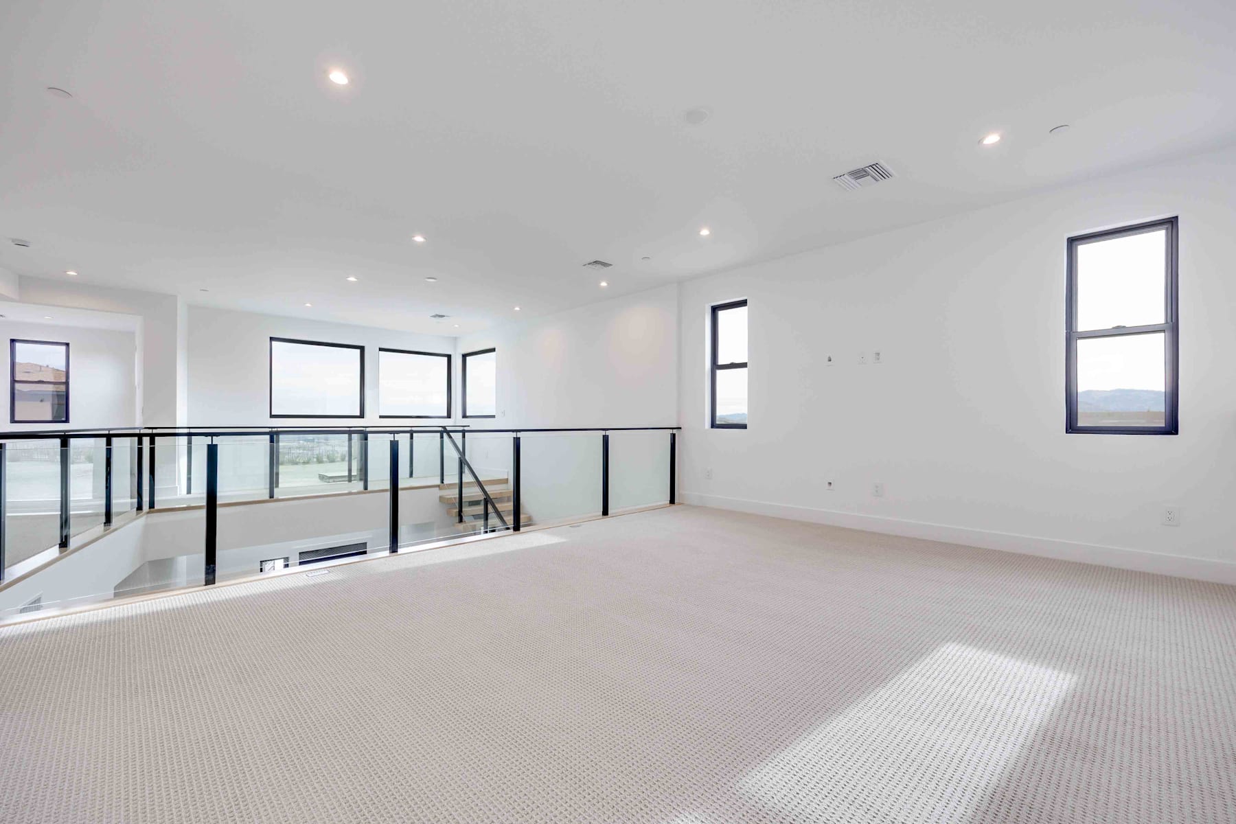 A spacious, well-lit room with a loft-style design, featuring a carpeted floor, large windows, and a glass railing overlooking the lower level.