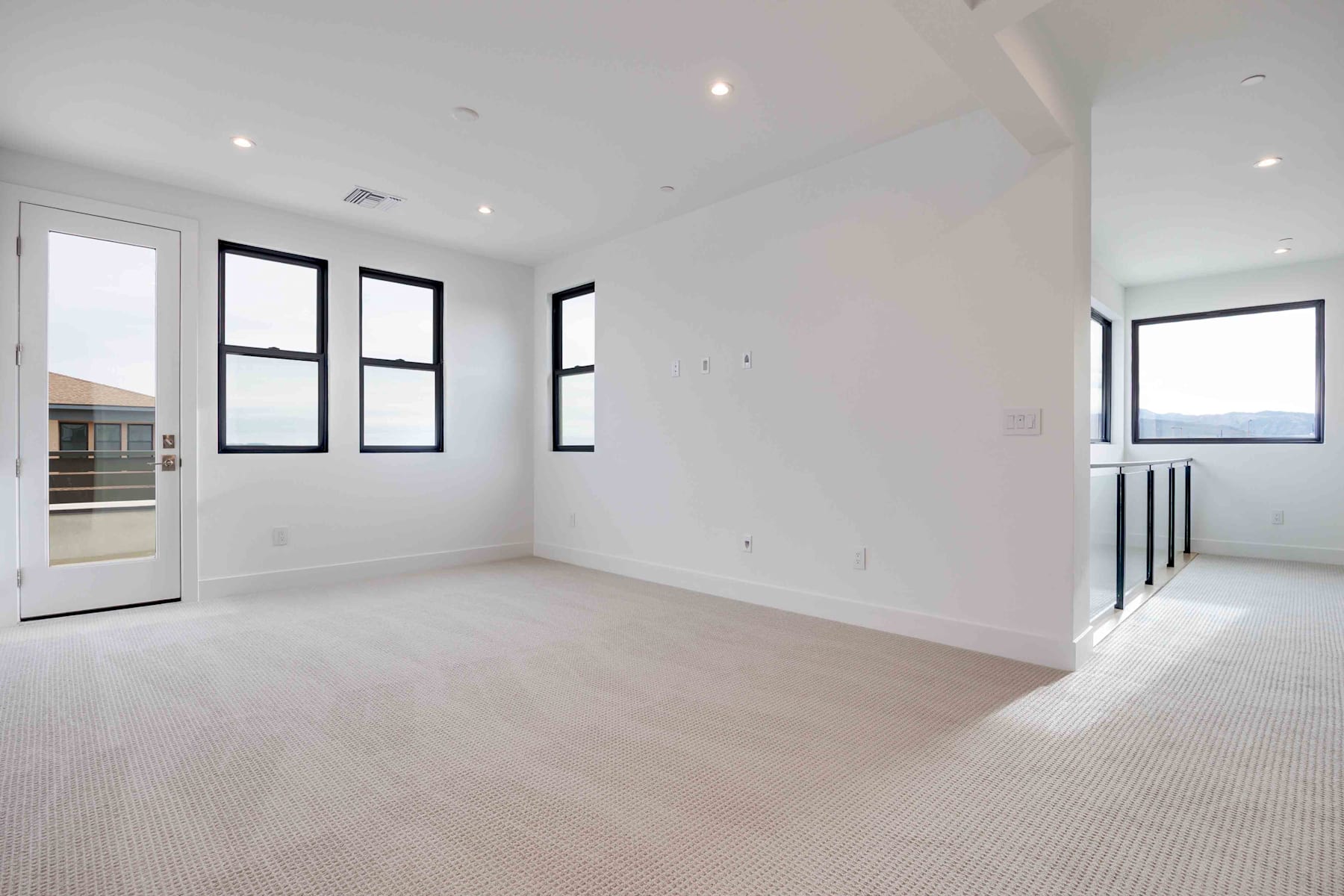 A spacious, well-lit room with white walls, large windows, and a carpeted floor. The room appears to be part of a modern, minimalist interior design.