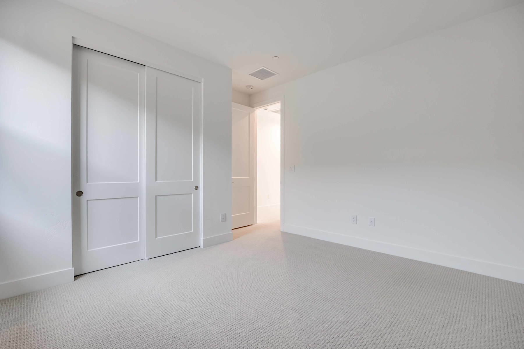 A simple, minimalist room with white walls, a closed door, and a carpeted floor, creating a clean and uncluttered atmosphere.