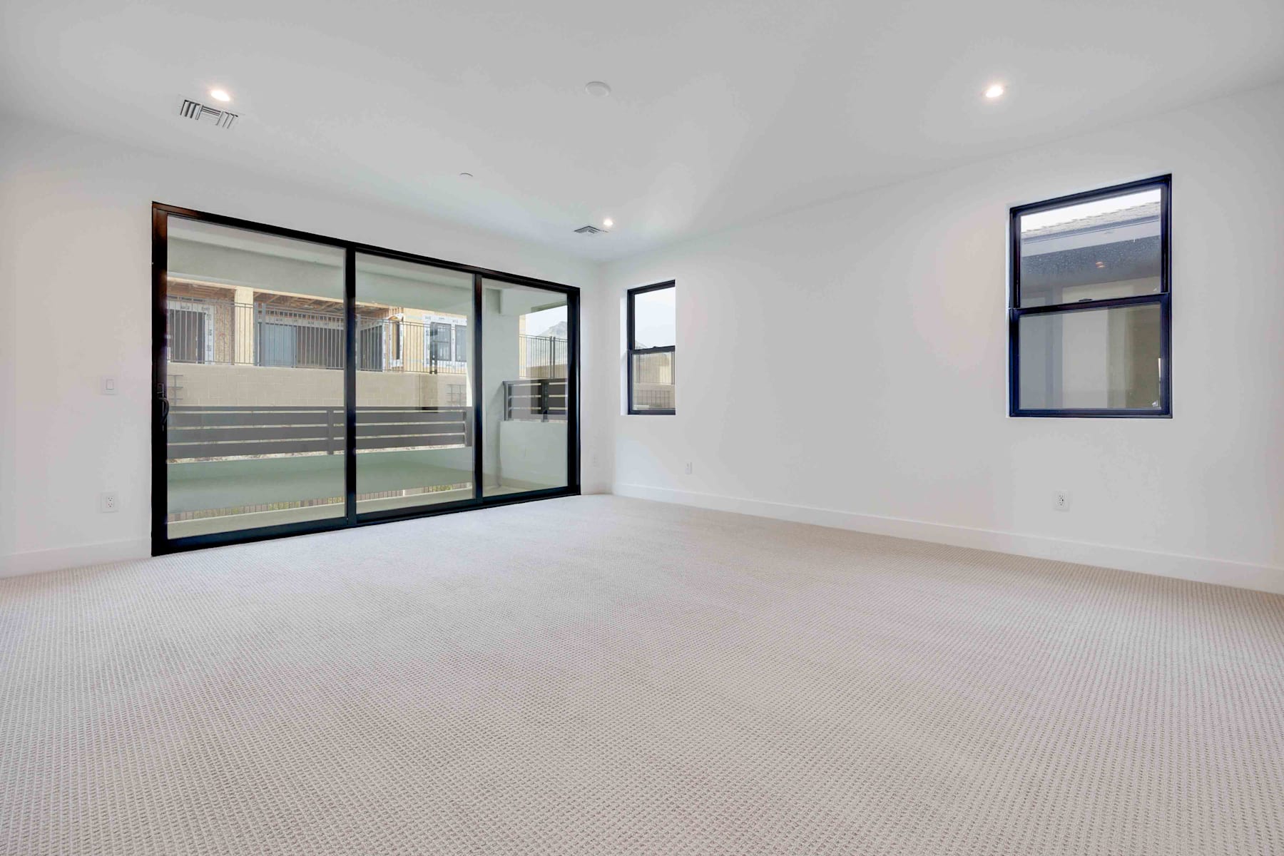 A spacious, minimalist room with large windows, white walls, and a carpeted floor. The room appears to be a living or bedroom space with a clean, modern aesthetic.
