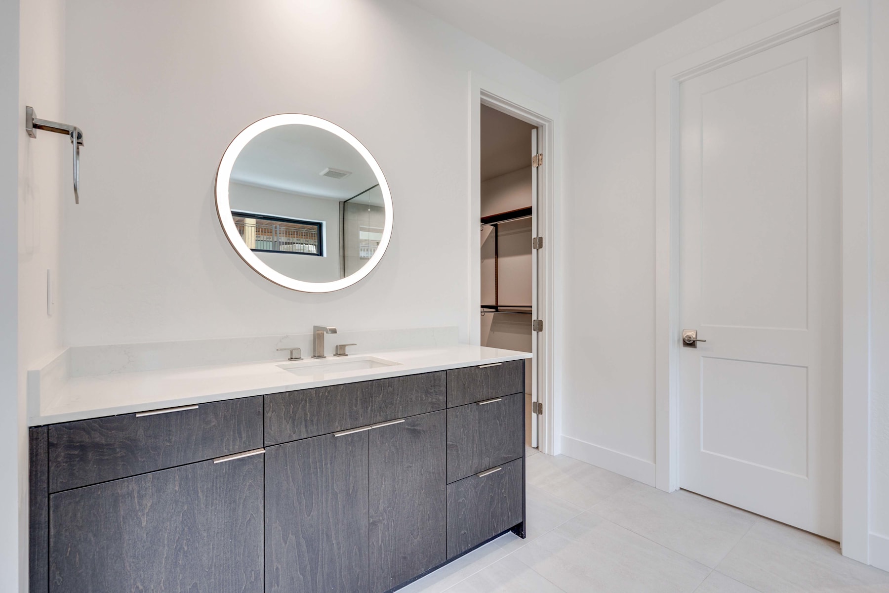 A modern bathroom with a large oval mirror, a dark wood vanity, and a doorway leading to another room.