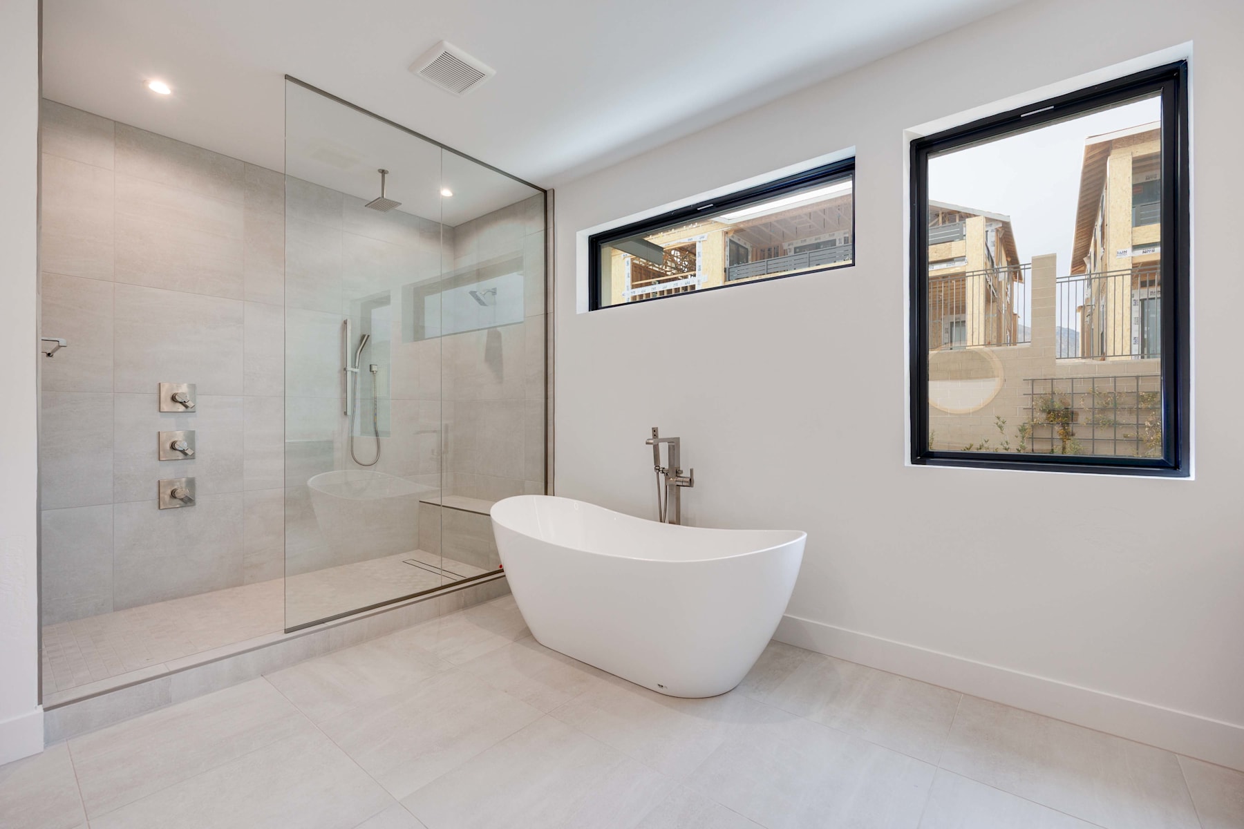 A modern, minimalist bathroom with a freestanding bathtub, glass shower enclosure, and large window providing natural light and a view of the exterior buildings.
