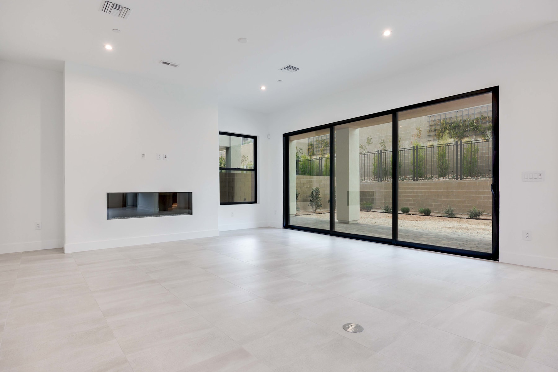A spacious, minimalist room with large glass windows overlooking a small outdoor area, featuring a fireplace and clean, white walls.