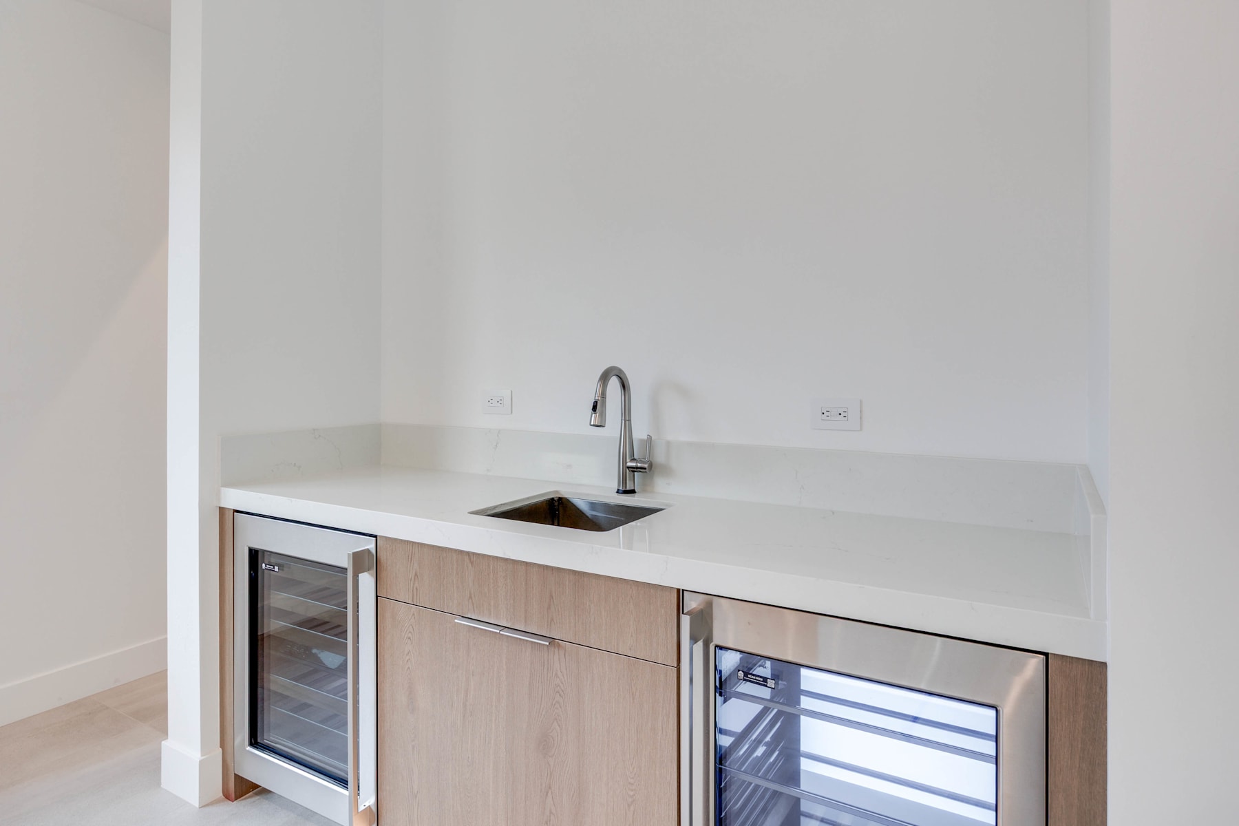 A modern kitchen with a white countertop, a stainless steel sink, and wooden cabinets, set against a plain white wall.