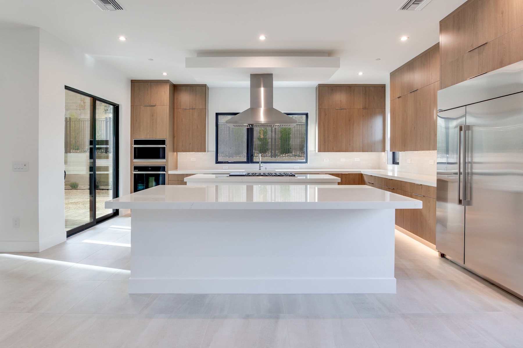 A modern, open-concept kitchen with a large white island, wooden cabinetry, and stainless steel appliances, set against a bright and airy backdrop.