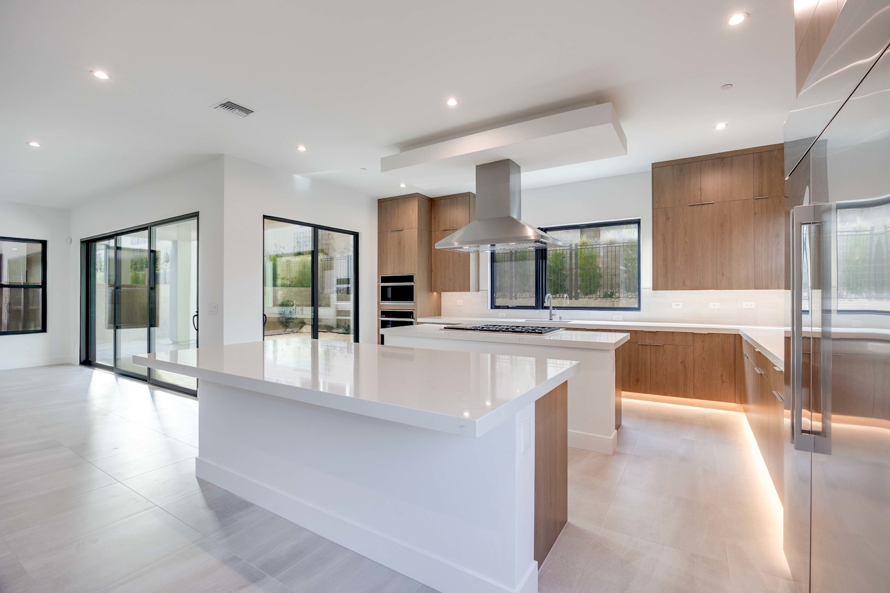A modern, open-concept kitchen with sleek white countertops, wooden cabinetry, and large windows overlooking a scenic outdoor view.