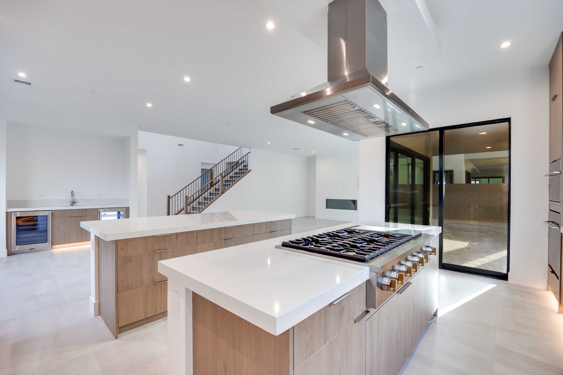 A modern, open-concept kitchen with a sleek, white countertop, a gas stove, and a copper-toned range hood, set against a backdrop of a bright, airy space with a staircase visible in the background.