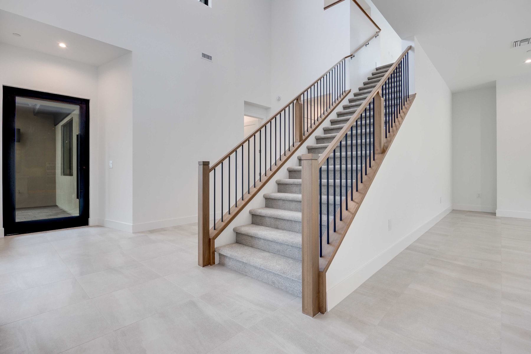 A modern, minimalist staircase with wooden handrails and metal balusters stands in a bright, open space with white walls and floors.