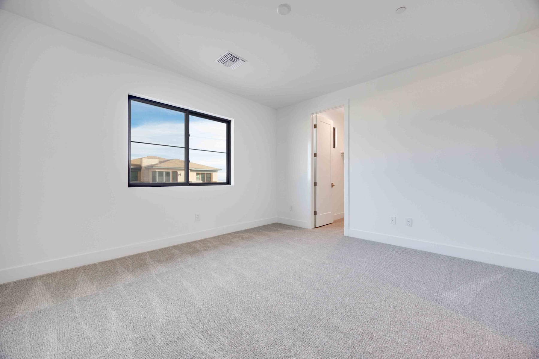 A spacious, well-lit room with a large window overlooking a building in the background, and a clean, minimalist interior with hardwood floors.