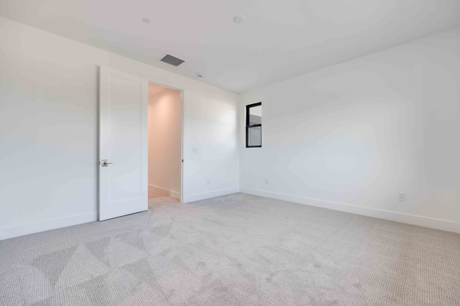 A minimalist, white-walled room with a carpeted floor and a closed door, creating a clean and uncluttered atmosphere.