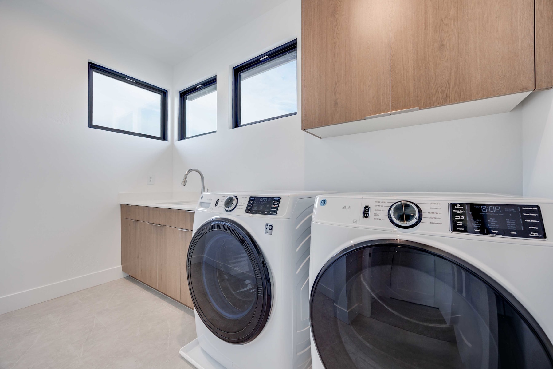 A modern and minimalist laundry room with a white washer and dryer set against a wooden cabinet backdrop, and large windows providing natural light.