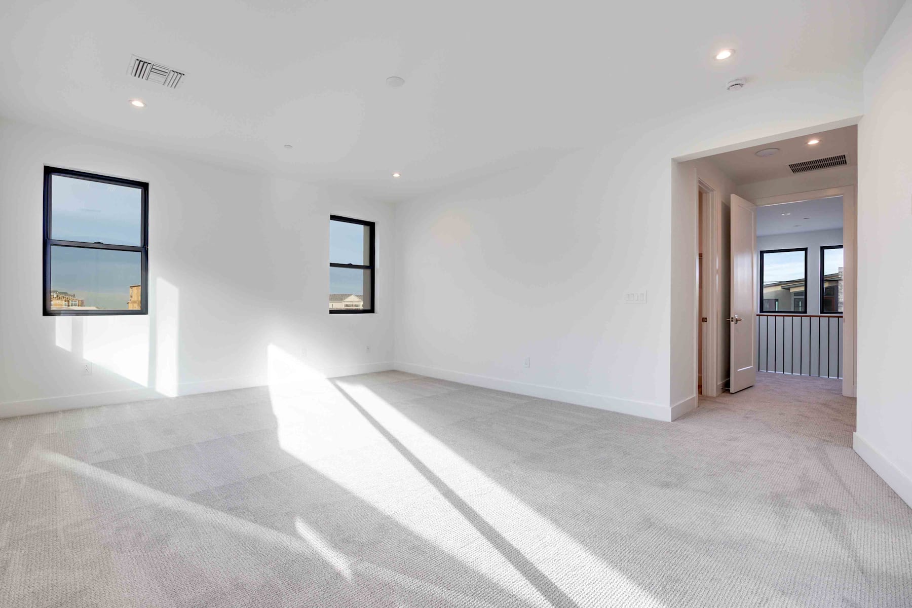 A spacious, well-lit room with white walls, a hardwood floor, and large windows providing a view of the exterior.