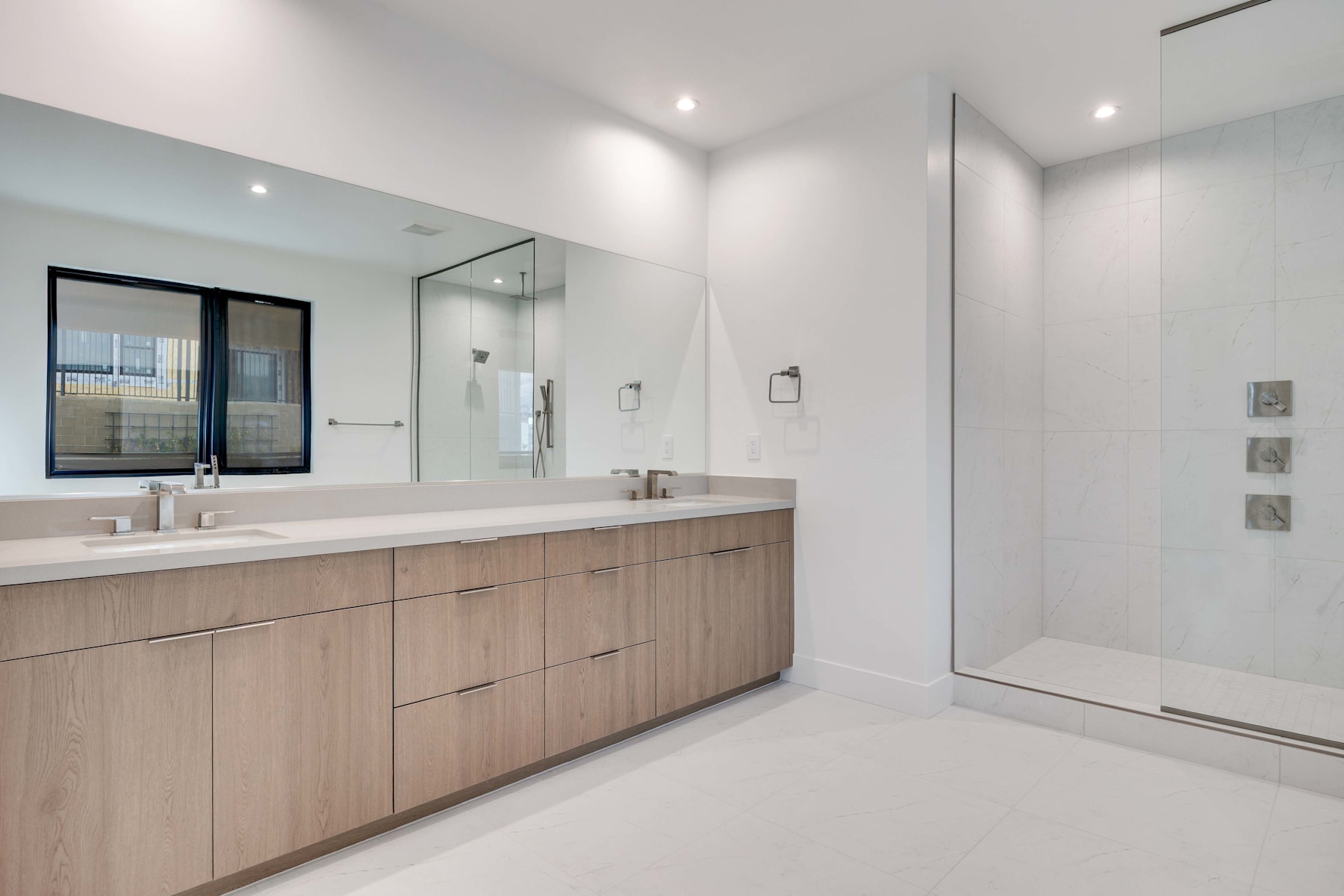 A modern and minimalist bathroom with a large vanity, a glass shower enclosure, and recessed lighting creating a bright and airy atmosphere.