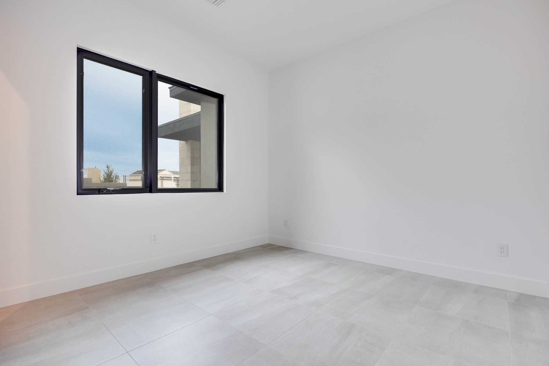 A minimalist, well-lit room with a large window framed by black metal, showcasing a plain white wall and a smooth concrete floor.