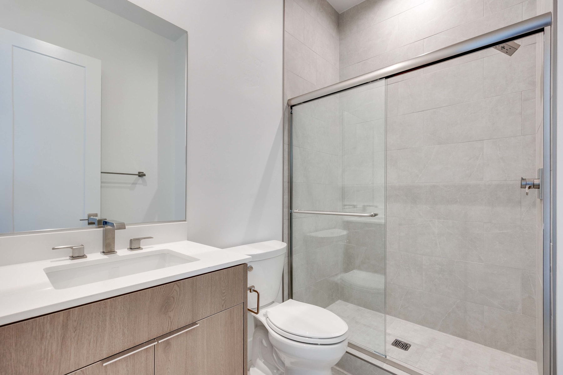 A modern bathroom with a vanity, sink, and a shower enclosure with a glass door, set against a neutral-toned tiled wall.