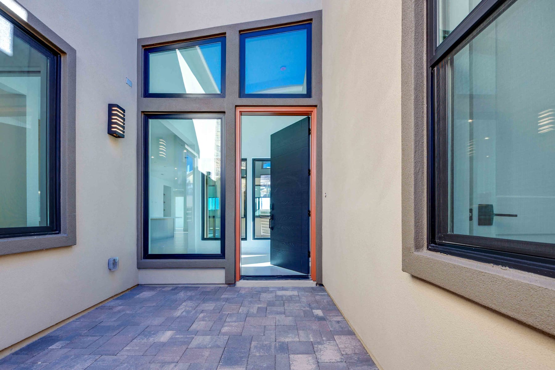 A modern, minimalist entryway with large glass windows and doors, leading to an outdoor area with a clear blue sky visible.