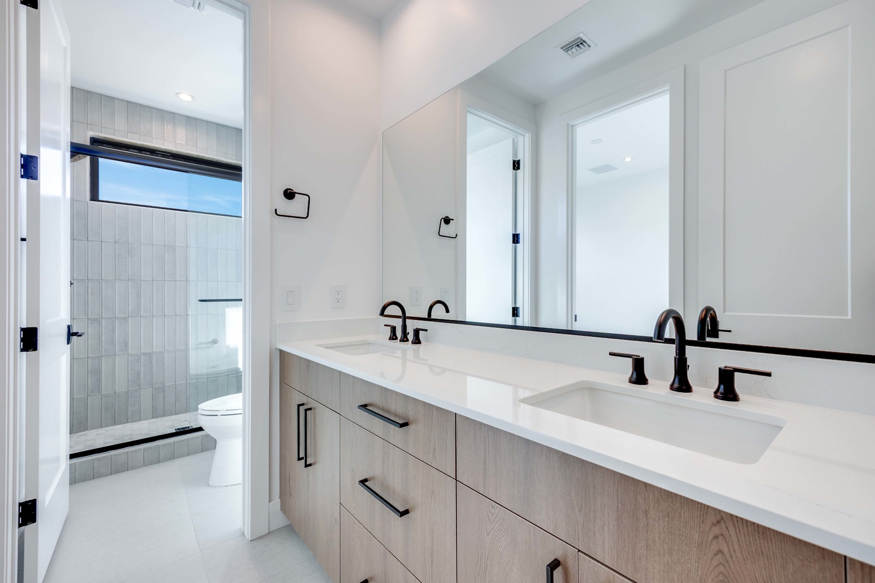 A modern bathroom with a double vanity, white countertop, and black faucets, set against a backdrop of light-colored walls and a glass shower enclosure.
