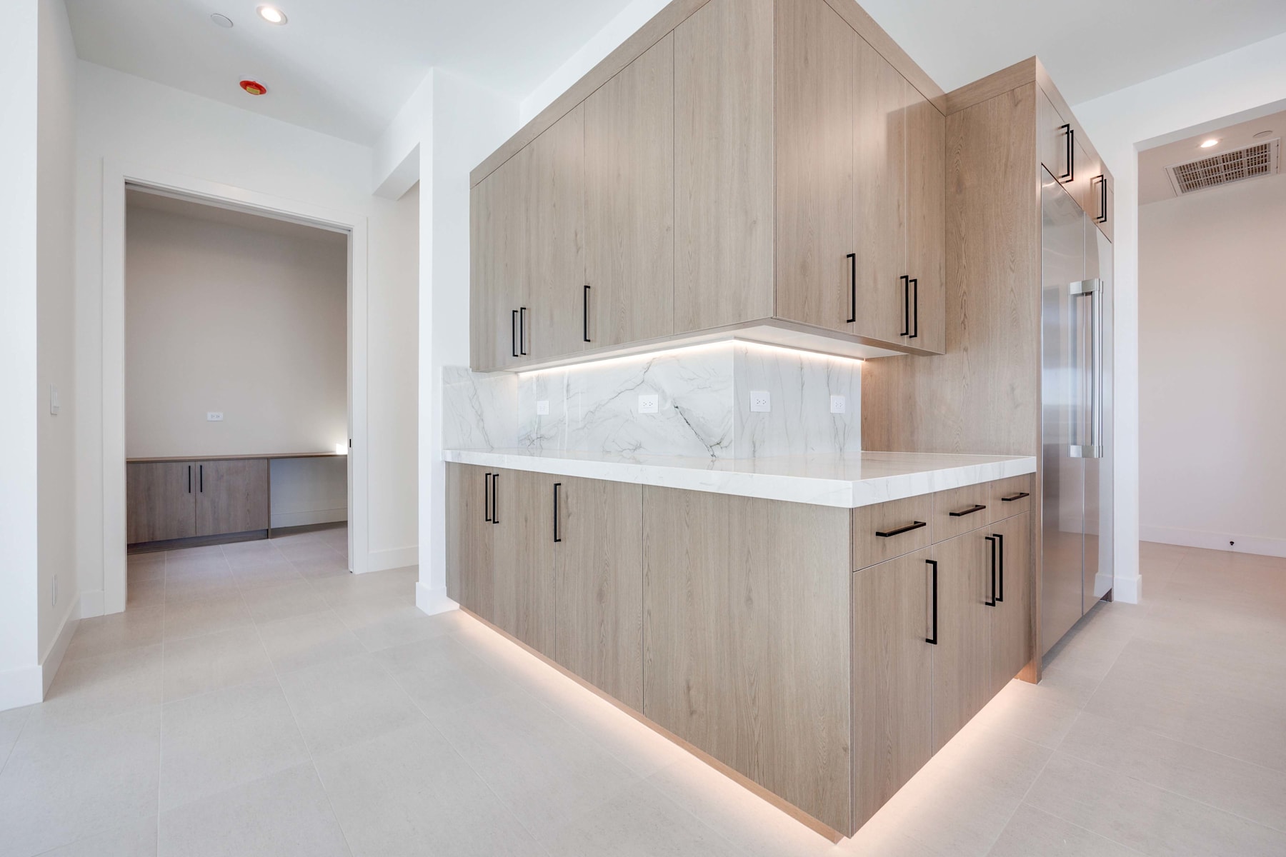 A modern, minimalist kitchen with light wood cabinets, a white marble countertop, and recessed lighting, set against a bright, open hallway.