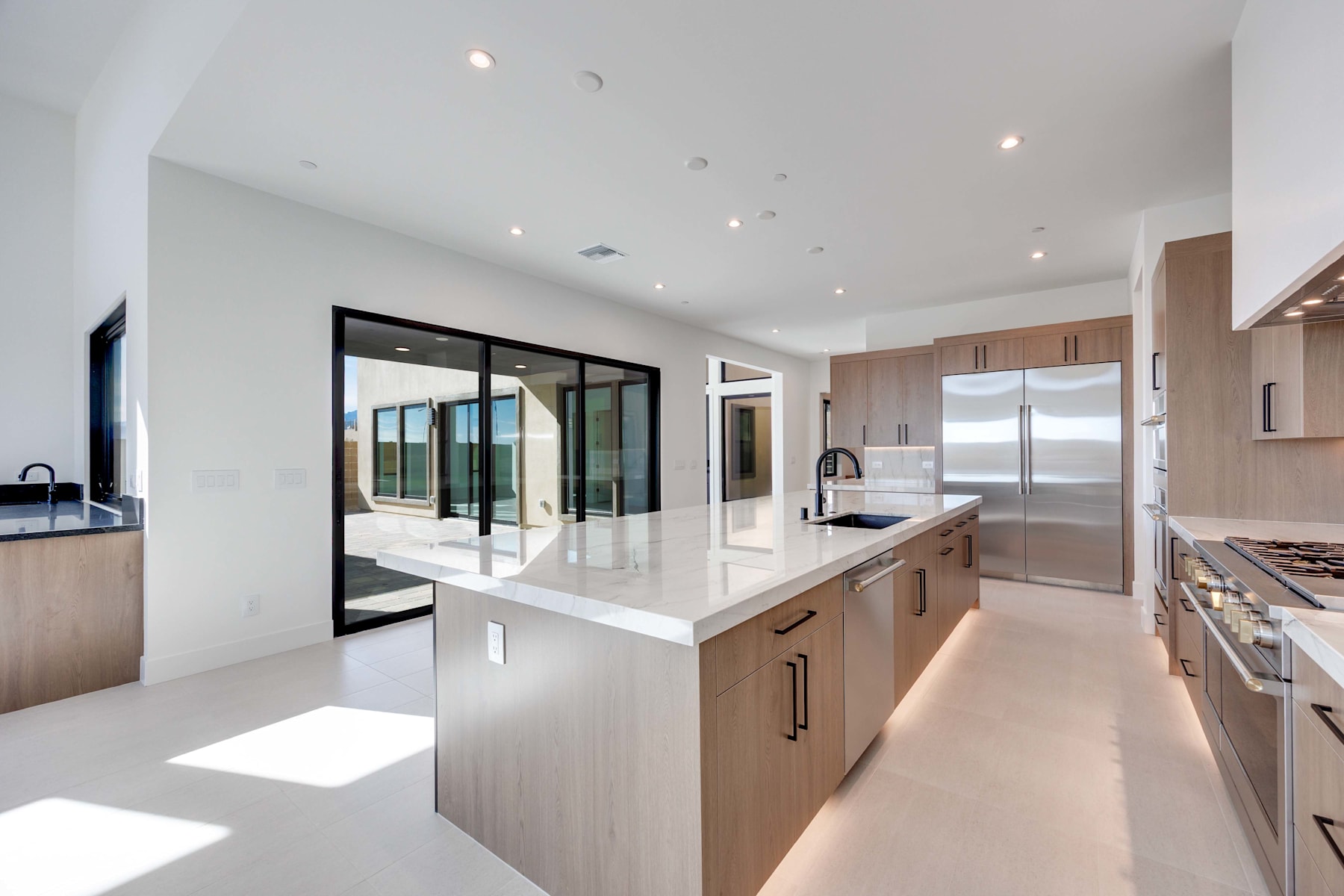 A modern, open-concept kitchen with light-colored cabinets, a large island with a white countertop, and a view of a living room area through glass doors.