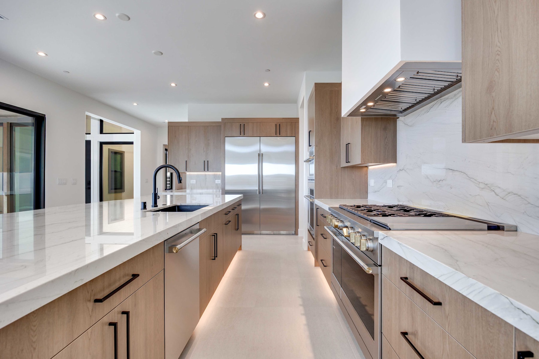 A modern, well-lit kitchen with sleek, light-colored cabinetry, stainless steel appliances, and a marble-like countertop that extends into the background, creating a spacious and visually appealing interior.