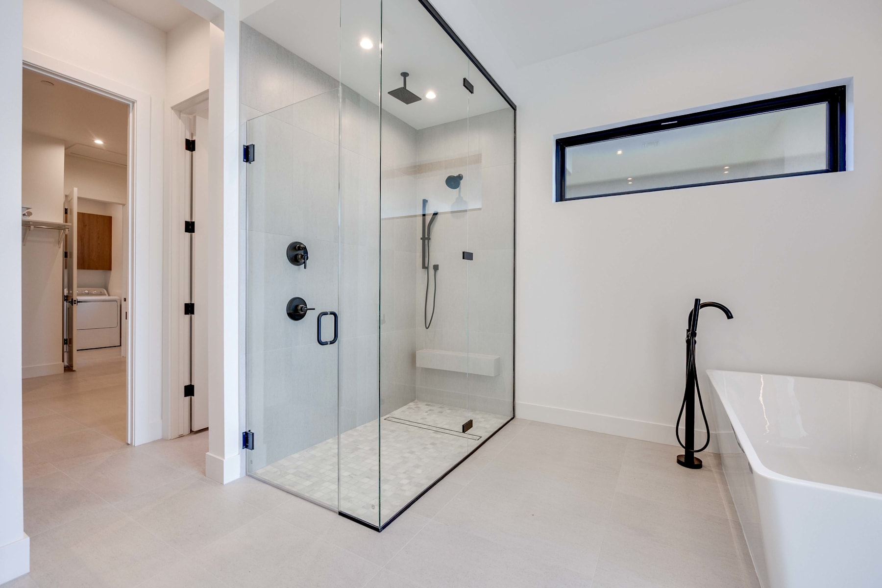 A modern, minimalist bathroom with a large glass shower enclosure, a freestanding bathtub, and a long horizontal window providing natural light.