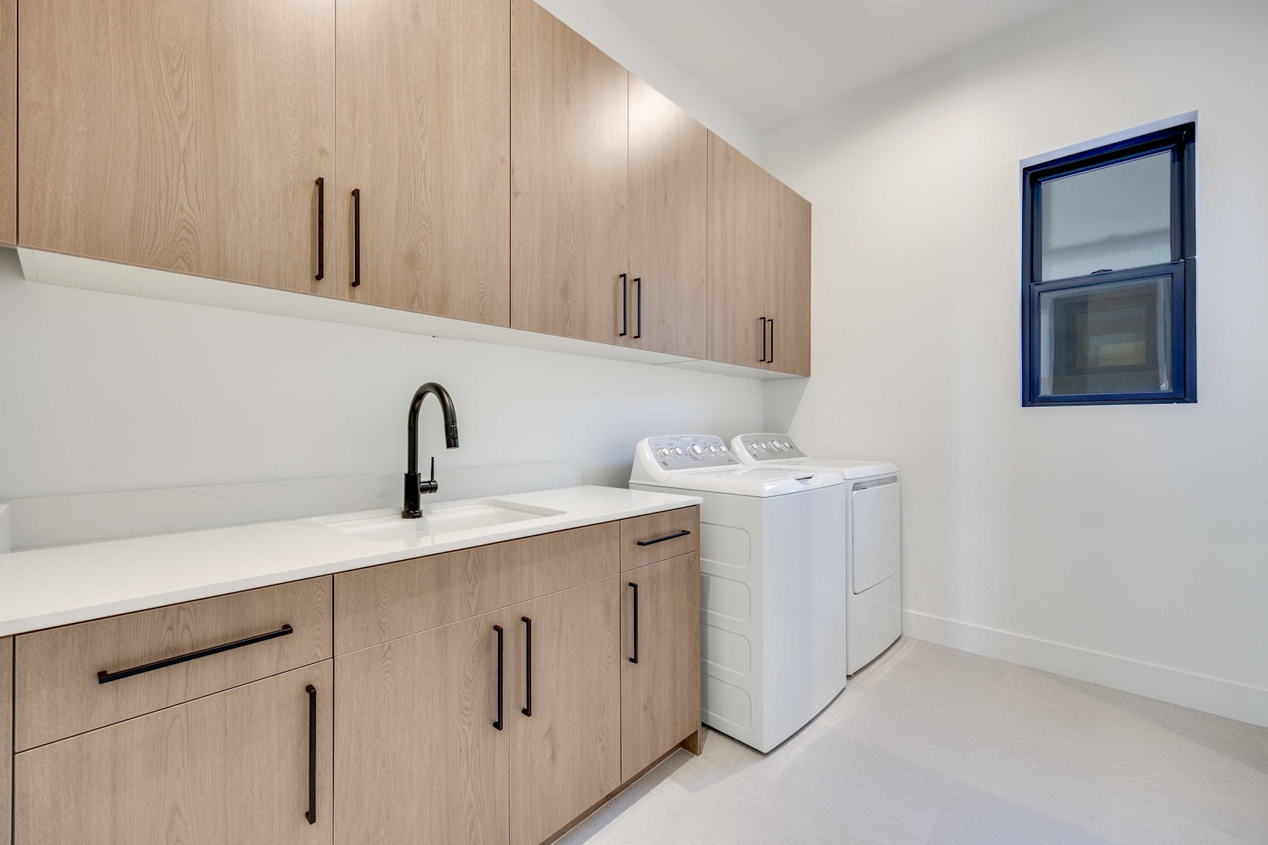 A modern and minimalist laundry room with light wood cabinets, a white countertop, and a washing machine and dryer set against a white wall.