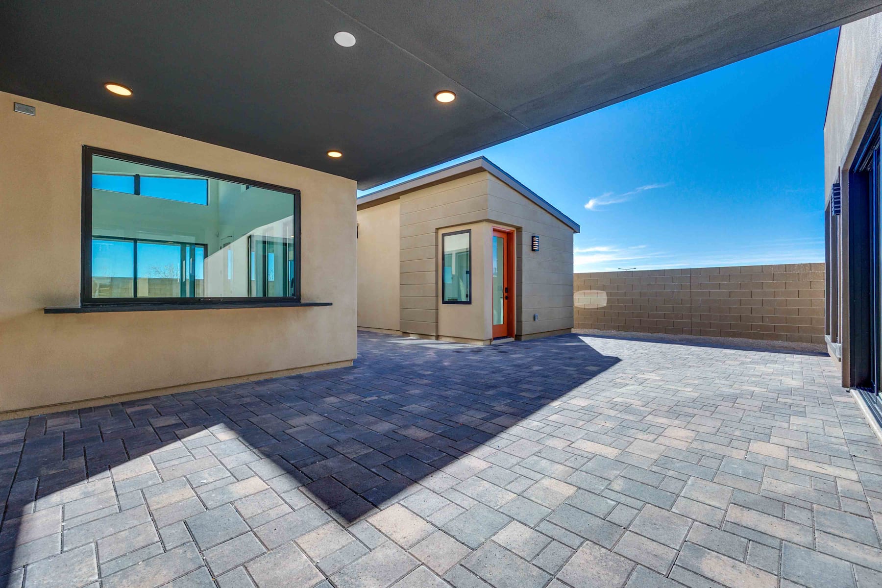A modern, well-designed exterior of a residential building with a paved courtyard, large windows, and a red door, set against a clear blue sky.