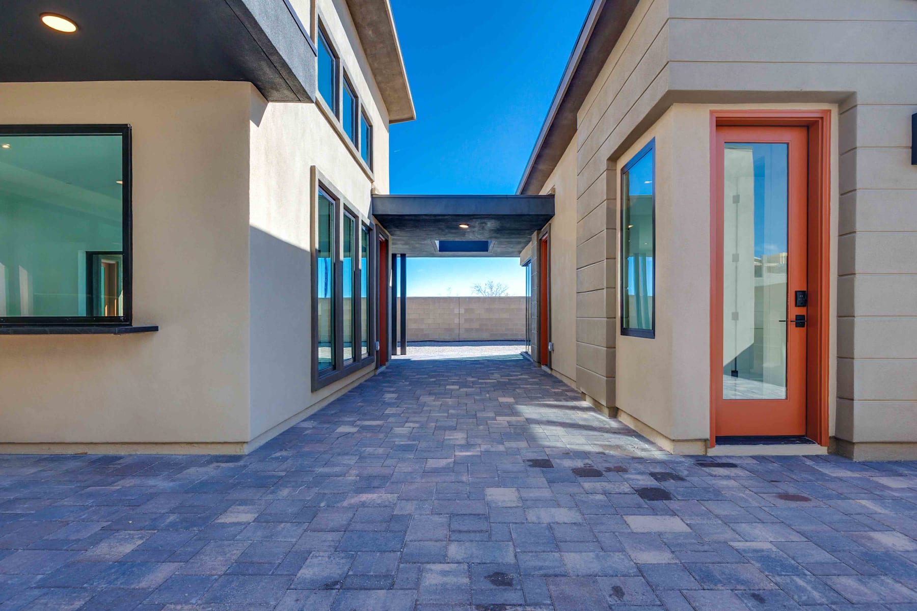 A paved walkway leads through a modern building with a vibrant orange door, surrounded by a clear blue sky.