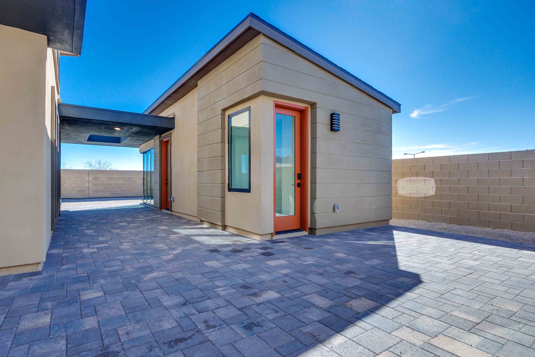 A modern, single-story house with a paved courtyard in the foreground and a clear blue sky in the background.