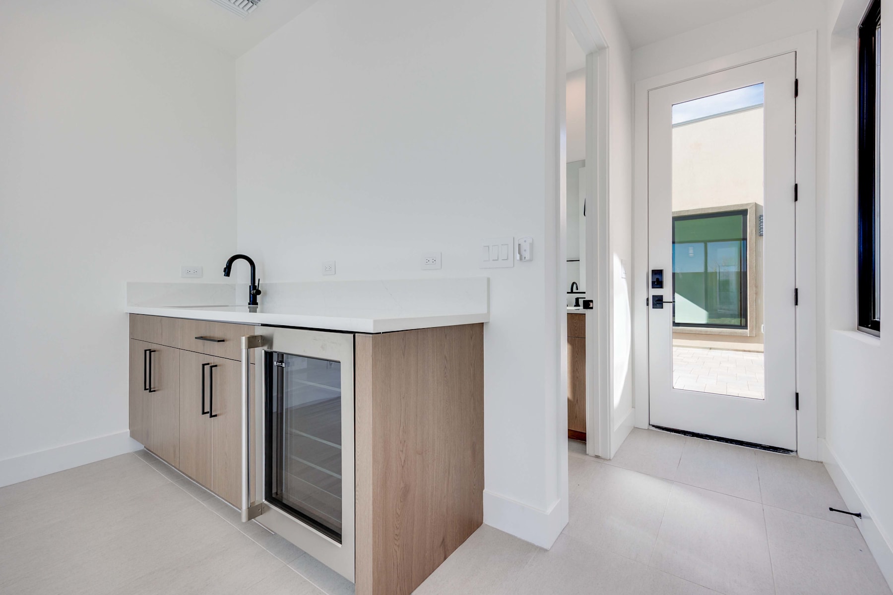 A modern, minimalist kitchen with a white countertop, wooden cabinets, and a glass-fronted refrigerator, set against a bright, airy background with a door leading to an exterior space.