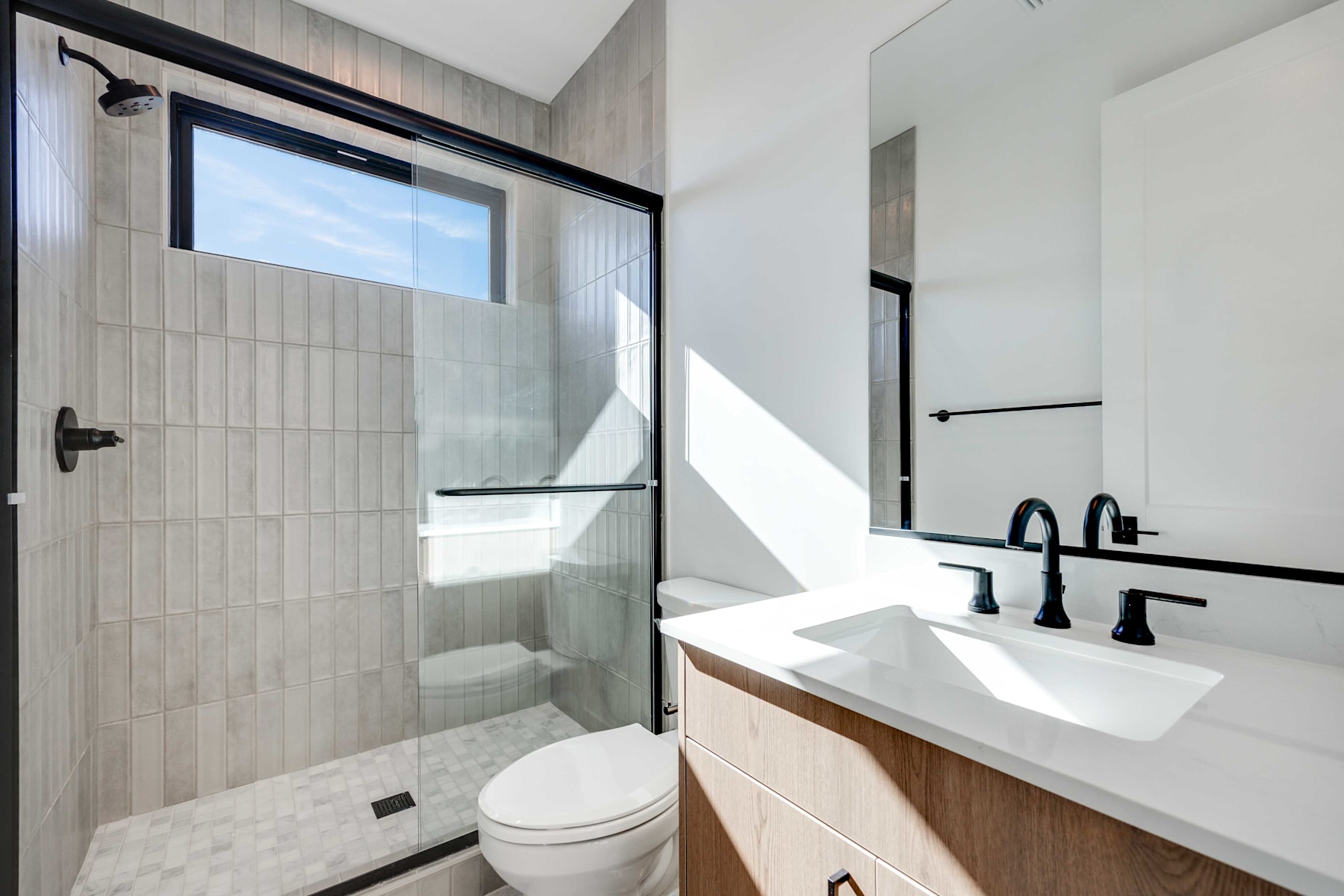 A modern bathroom with a glass-enclosed shower, a vanity with a sink, and a toilet, all set against a backdrop of light-colored tiles and a window providing natural lighting.