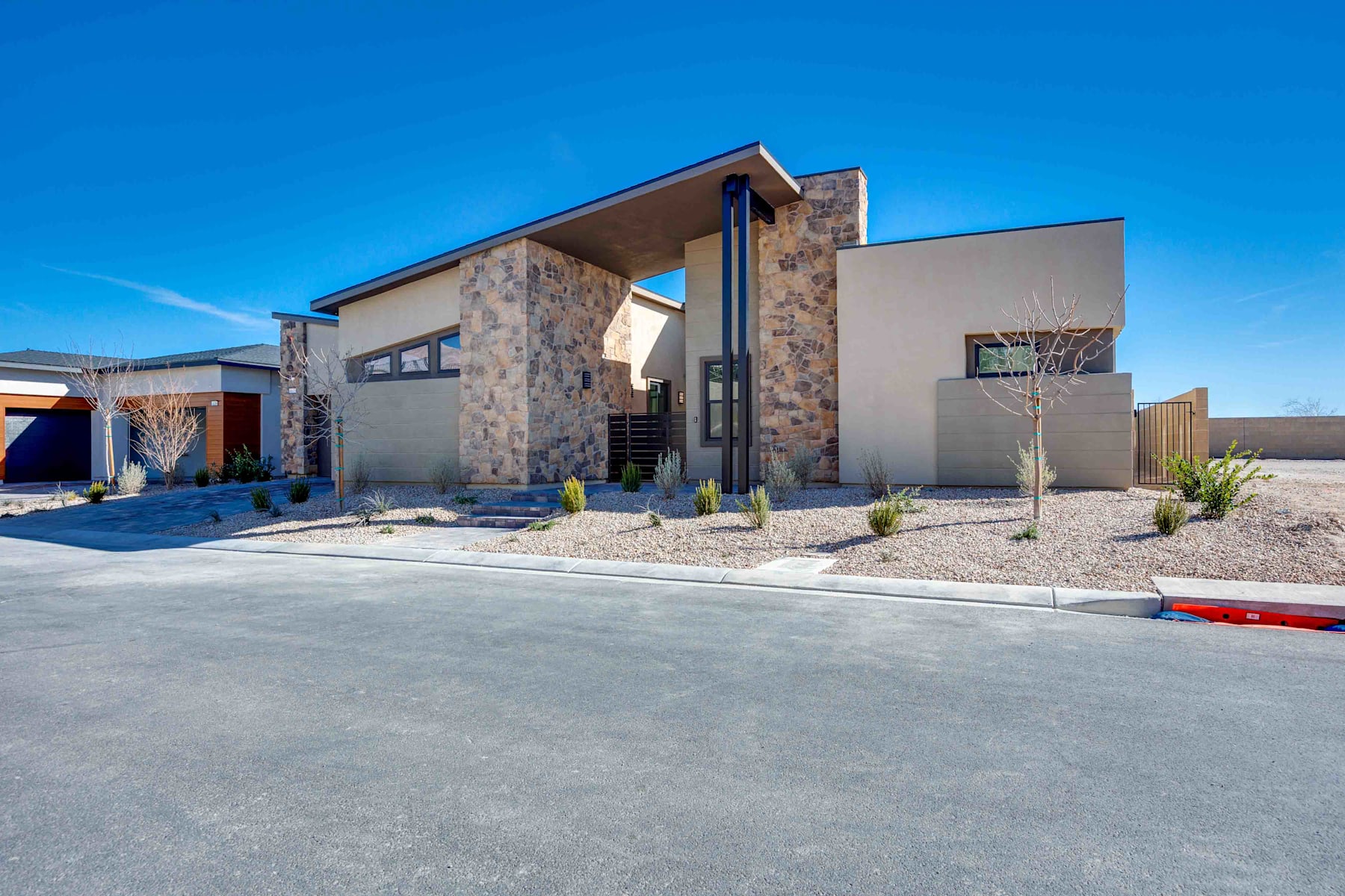 A modern, two-story residential building with a mix of stone and stucco exterior, surrounded by a paved driveway and desert landscaping against a clear blue sky.