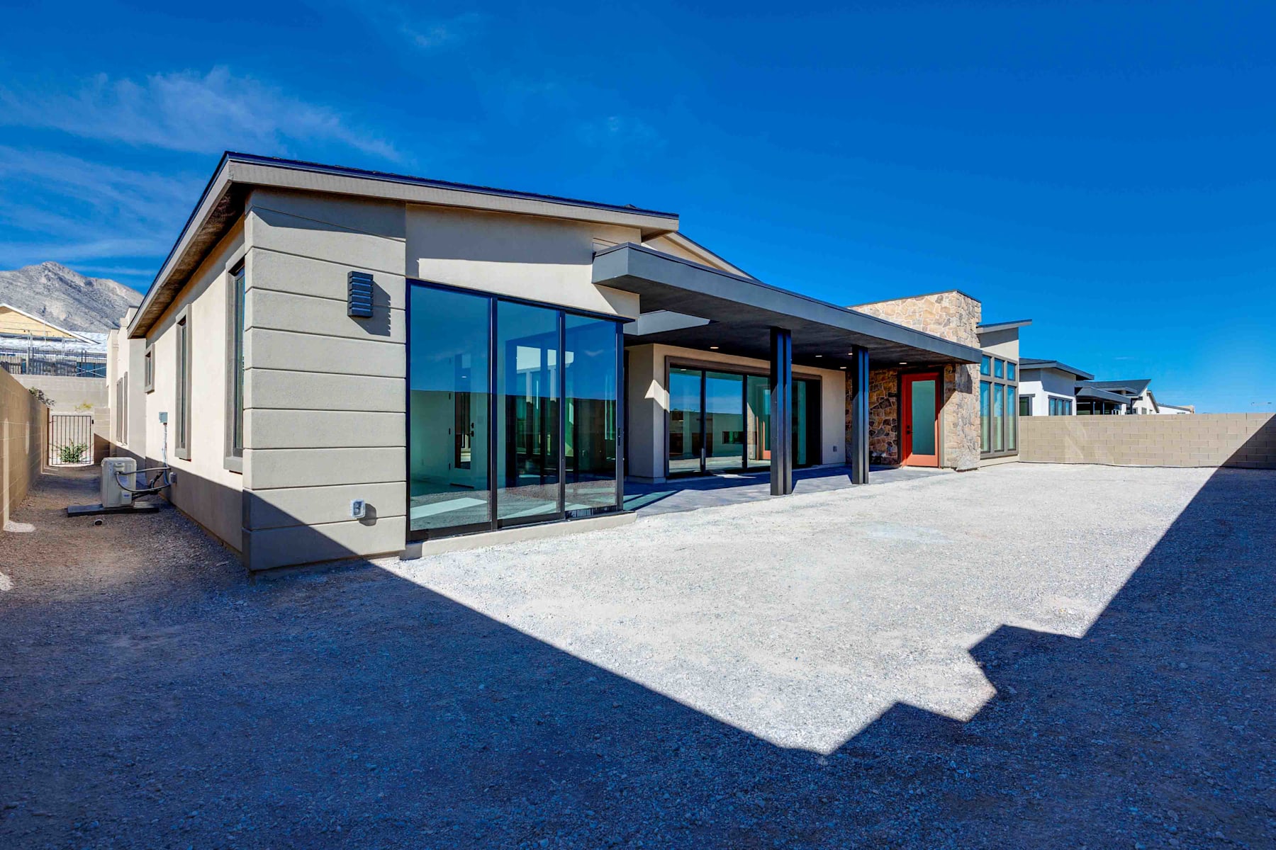 A modern, glass-fronted building with a flat roof stands in the foreground, surrounded by a paved area and set against a clear blue sky in the background.