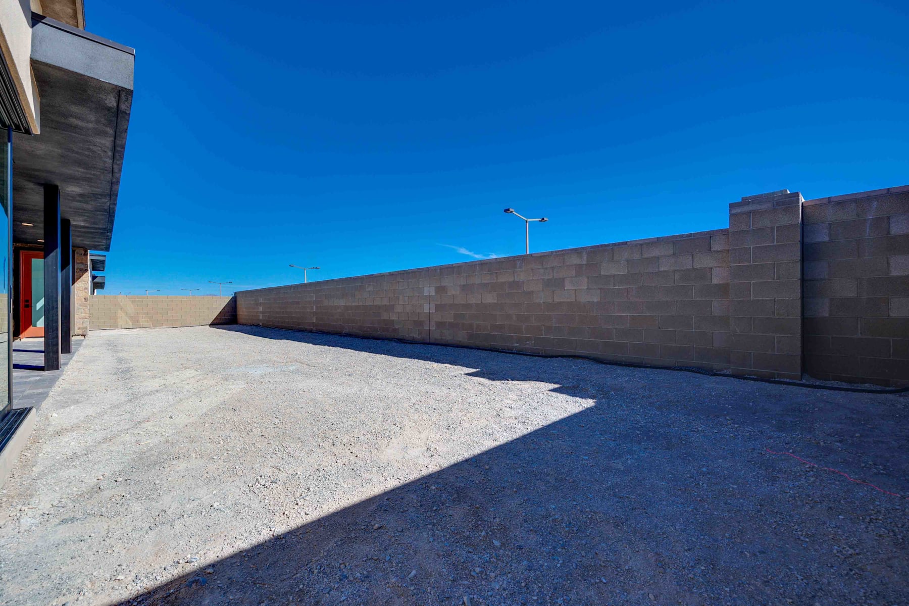 A paved walkway leads towards a tall concrete wall under a bright blue sky, with a street light visible in the distance.