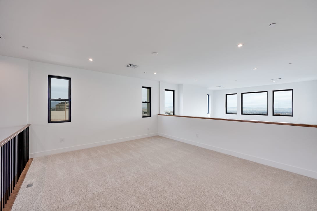 A spacious, well-lit room with white walls, large windows, and a carpeted floor. The room appears to be a living or bedroom area, with a railing visible in the background.