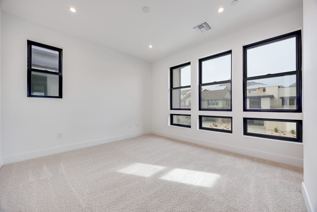 A bright, spacious room with large windows overlooking a residential area, featuring a clean, minimalist design with white walls and a plush, light-colored carpet.
