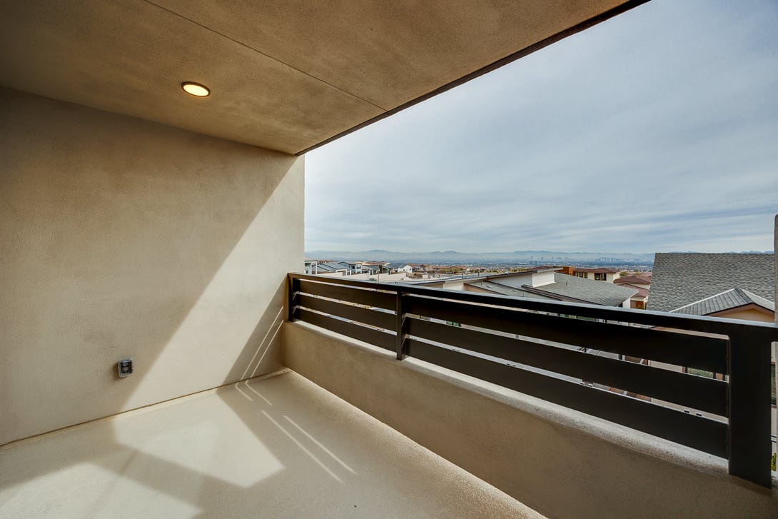 A balcony with a wooden railing overlooks a cityscape in the distance, with a cloudy sky visible above.
