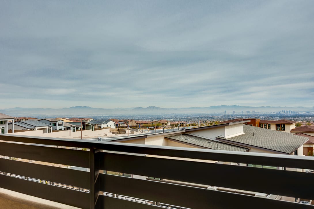 The image shows a cityscape with a view of rooftops and buildings in the foreground, with mountains visible in the background under a cloudy sky.