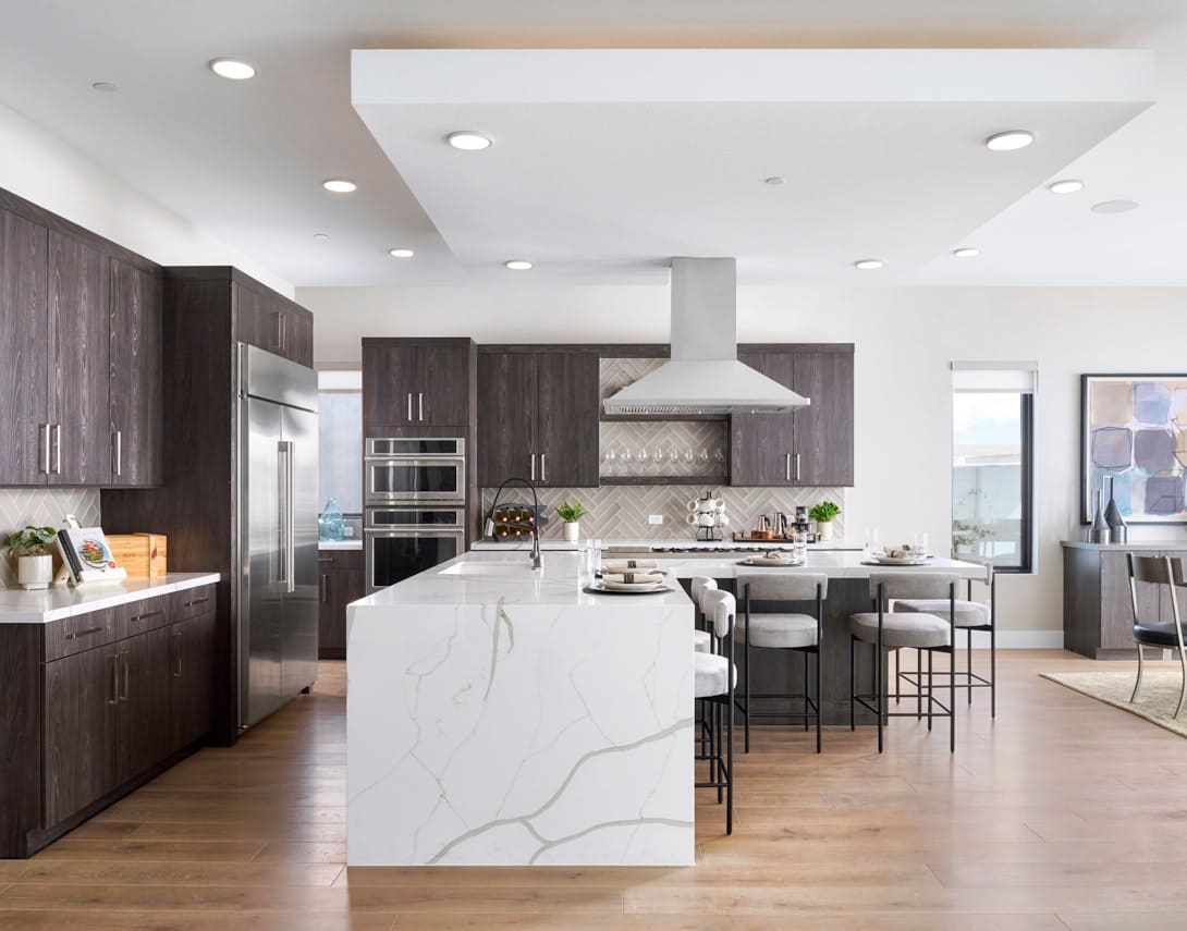 A modern, open-concept kitchen with dark wood cabinets, a large white marble island, and stainless steel appliances, set against a bright and airy background.