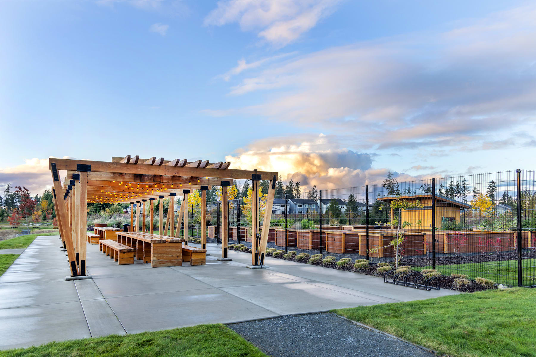 A wooden pergola structure with benches and planters stands in the foreground, overlooking a grassy area with trees and a cloudy sky in the background.