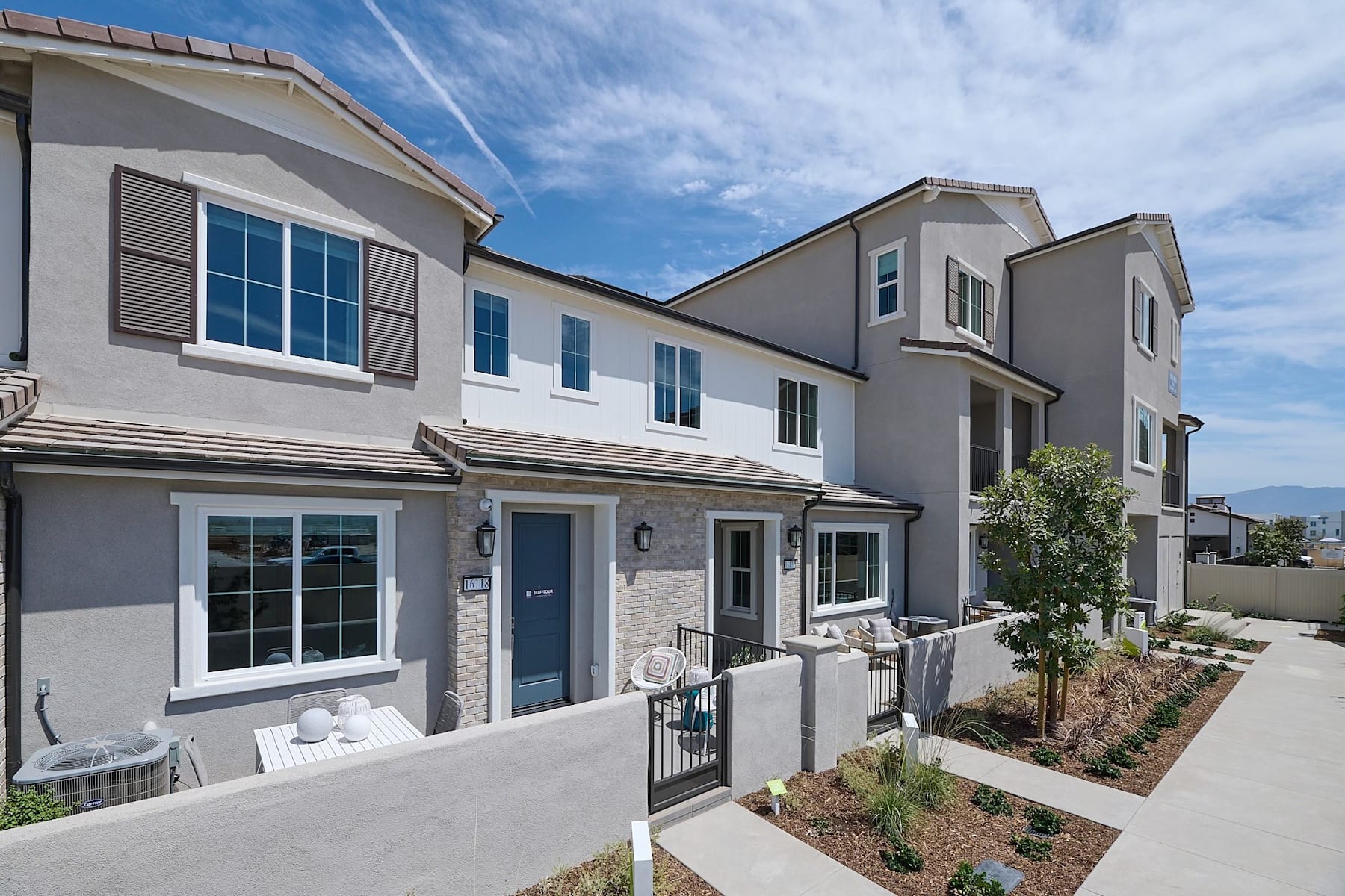 A modern, multi-unit residential building with a gray exterior, blue accents, and a well-landscaped front yard against a backdrop of a clear blue sky.