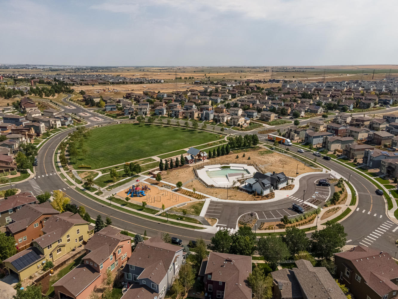 A sprawling residential neighborhood with a central park and circular road system, surrounded by rolling hills and open fields in the distance.