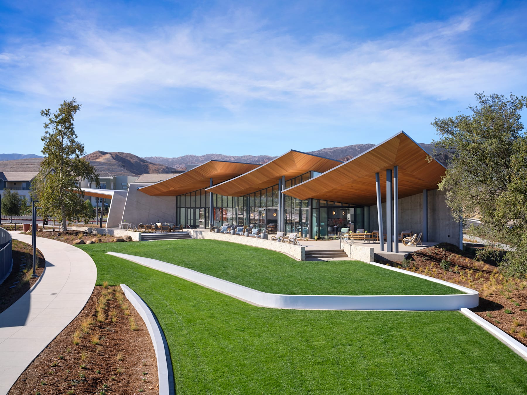 A modern, architecturally-designed building with a curved, orange-colored roof stands in the foreground, surrounded by a well-manicured lawn and landscaping. In the background, a mountainous landscape can be seen under a clear, blue sky.