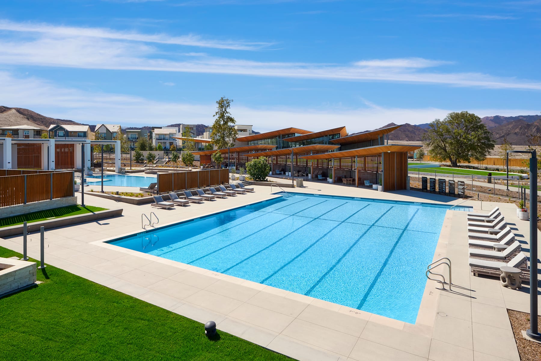 A large outdoor swimming pool surrounded by lounge chairs and umbrellas, with a backdrop of colorful buildings and a clear blue sky.