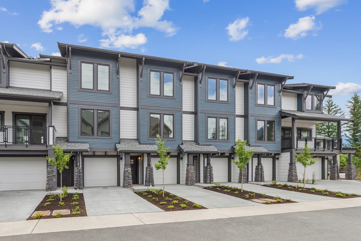 A row of modern, multi-story townhouses with gray siding and large windows, set against a backdrop of a blue sky with fluffy white clouds. The foreground features landscaped gardens and driveways leading to the attached garages.