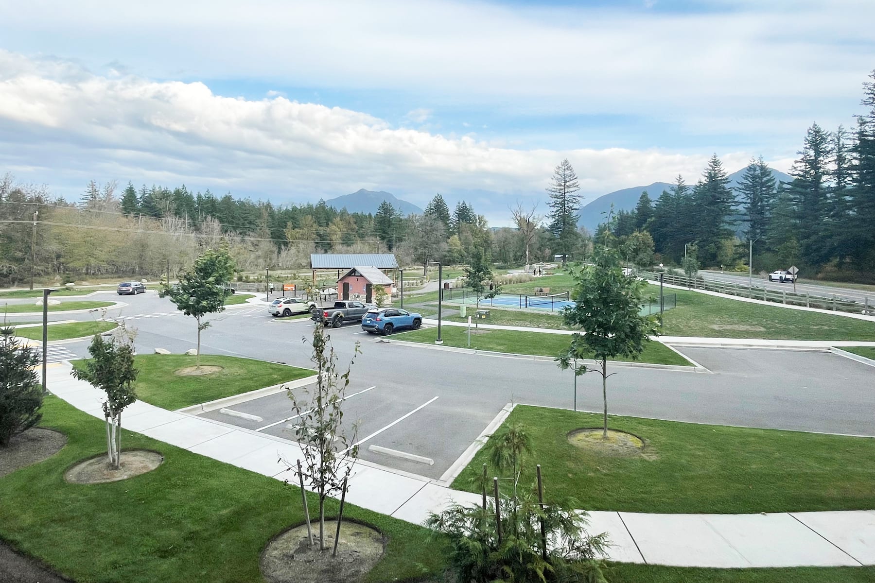 A peaceful suburban neighborhood with well-maintained lawns, trees, and roads, surrounded by a forested mountain landscape under a partly cloudy sky.