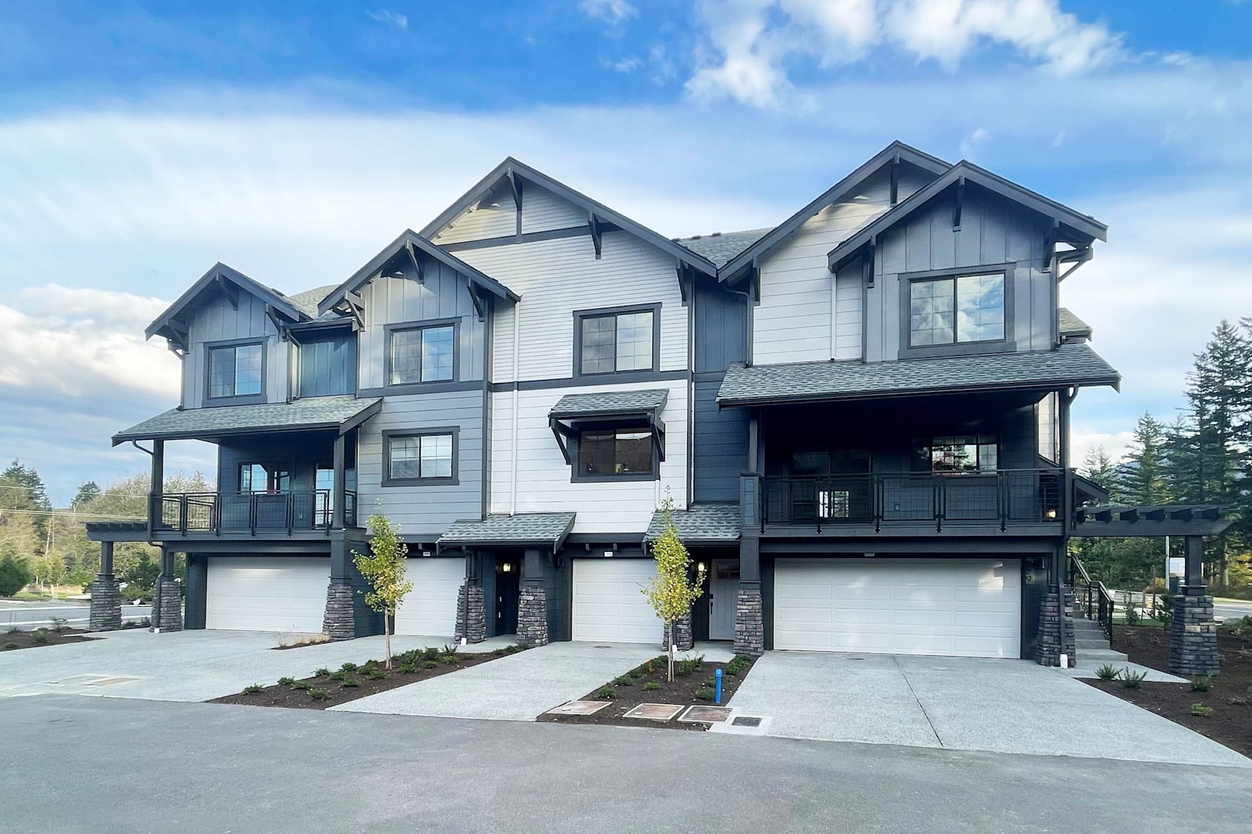 A modern, multi-unit townhouse complex with a mix of gray and white siding, gabled roofs, and attached garages, set against a backdrop of a blue sky with scattered clouds.