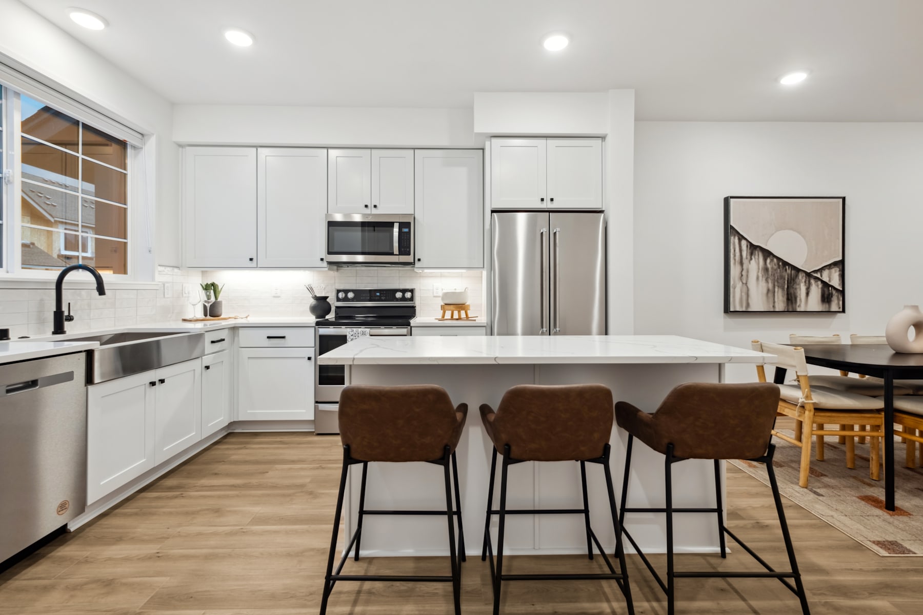 A modern, open-concept kitchen with white cabinets, stainless steel appliances, a large island with bar stools, and a framed artwork on the wall.