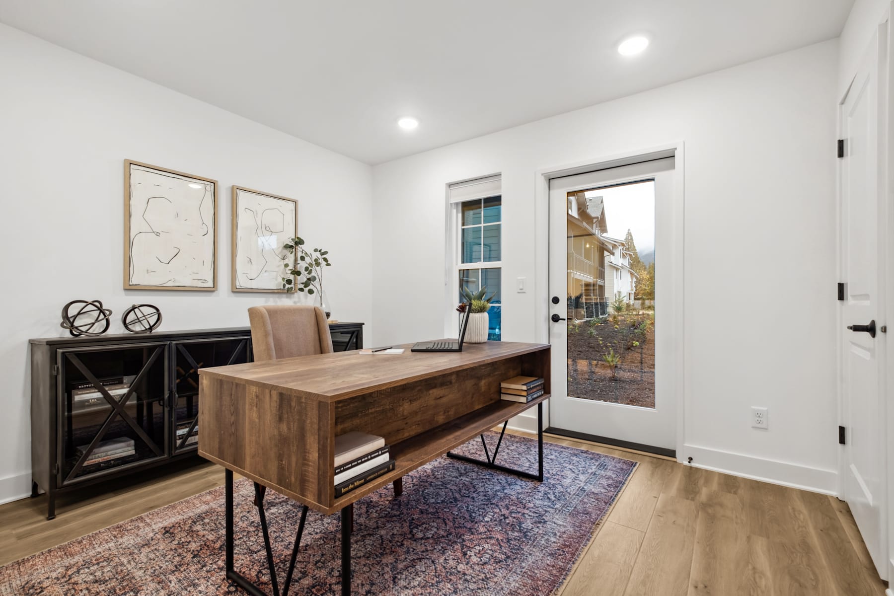 A modern and minimalist home office space with a wooden desk, shelving unit, and artwork on the walls, overlooking a scenic outdoor view through a glass door.